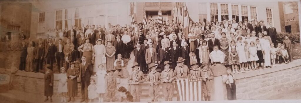 Four hundred men, women, and children stand in front of a high school in 1932. The wear clothing of the time, and the photo is black and white.