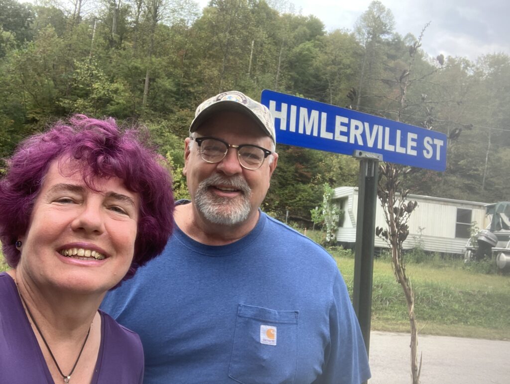 A women and man stand next to each other to take a selfie. She has purple hair, and he is wearing a baseball hat and glasses. Behind them is a street sign that says Himlerville Street.
