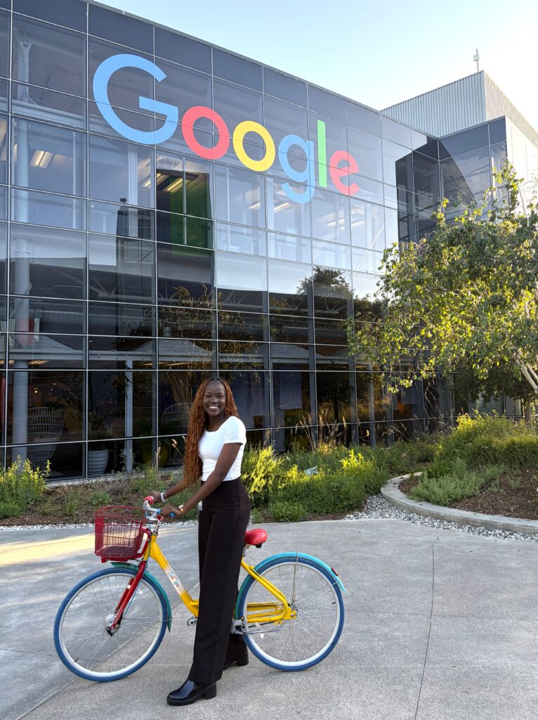 Female standing with her bike in front of a glass office building with the Google logo on it.