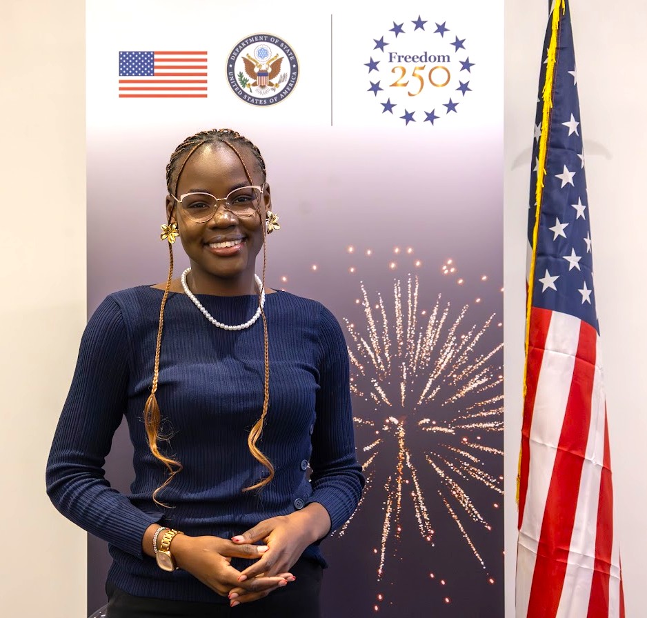 Female in her mid-twenties standing in front of an American flag and poster that has the America 250 and U.S. State Department logos and fireworks on it.
